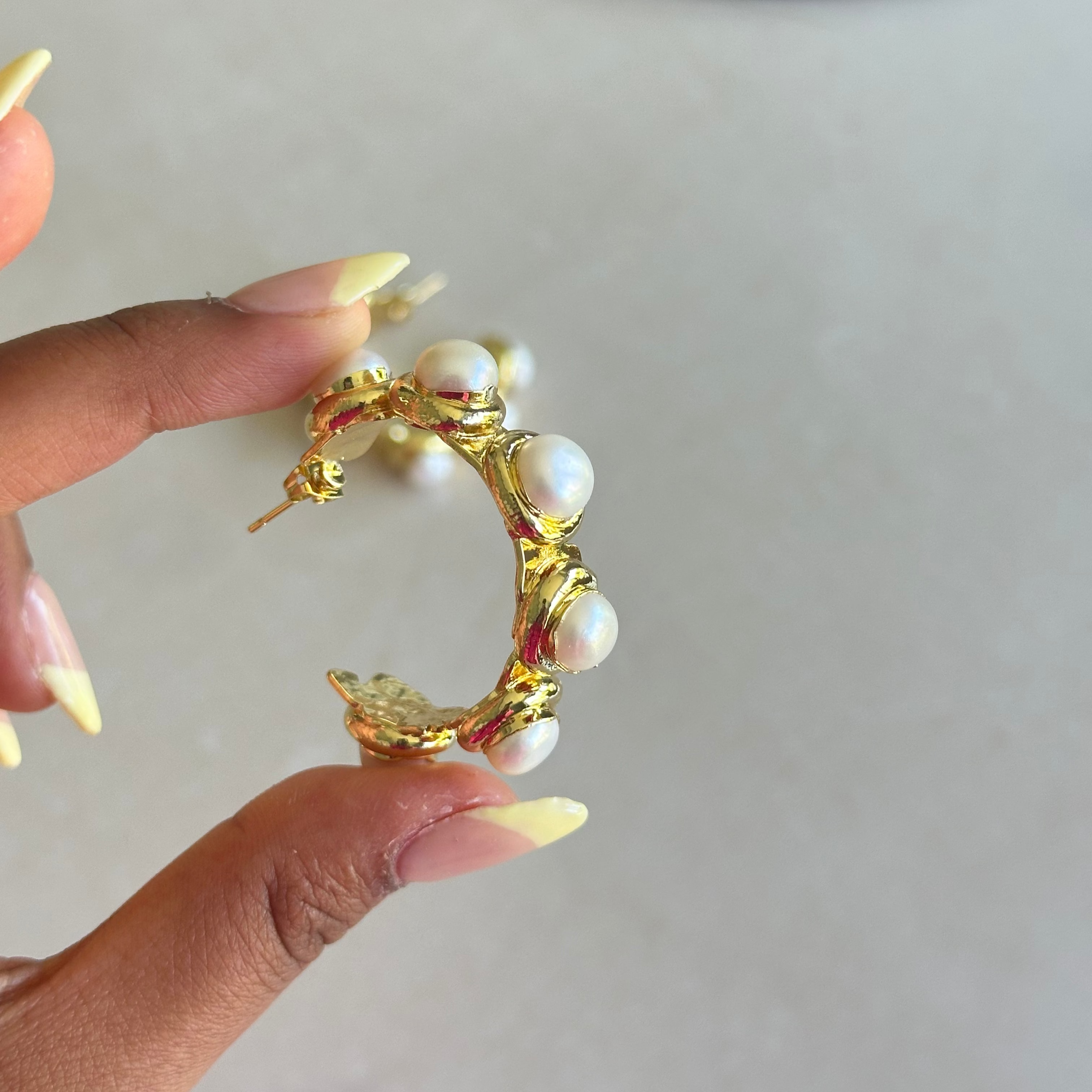 Gold hoop earring with pearls held by a hand against a neutral background